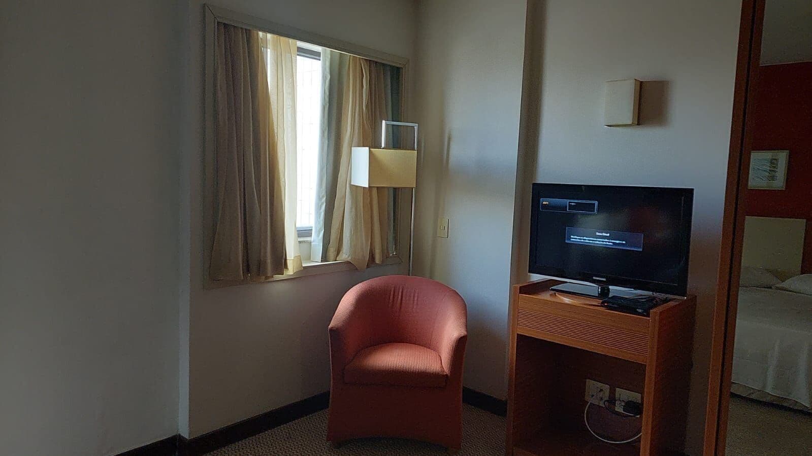 A hotel room with a single bed, a television on a wooden stand, a red chair, and a window with curtains.