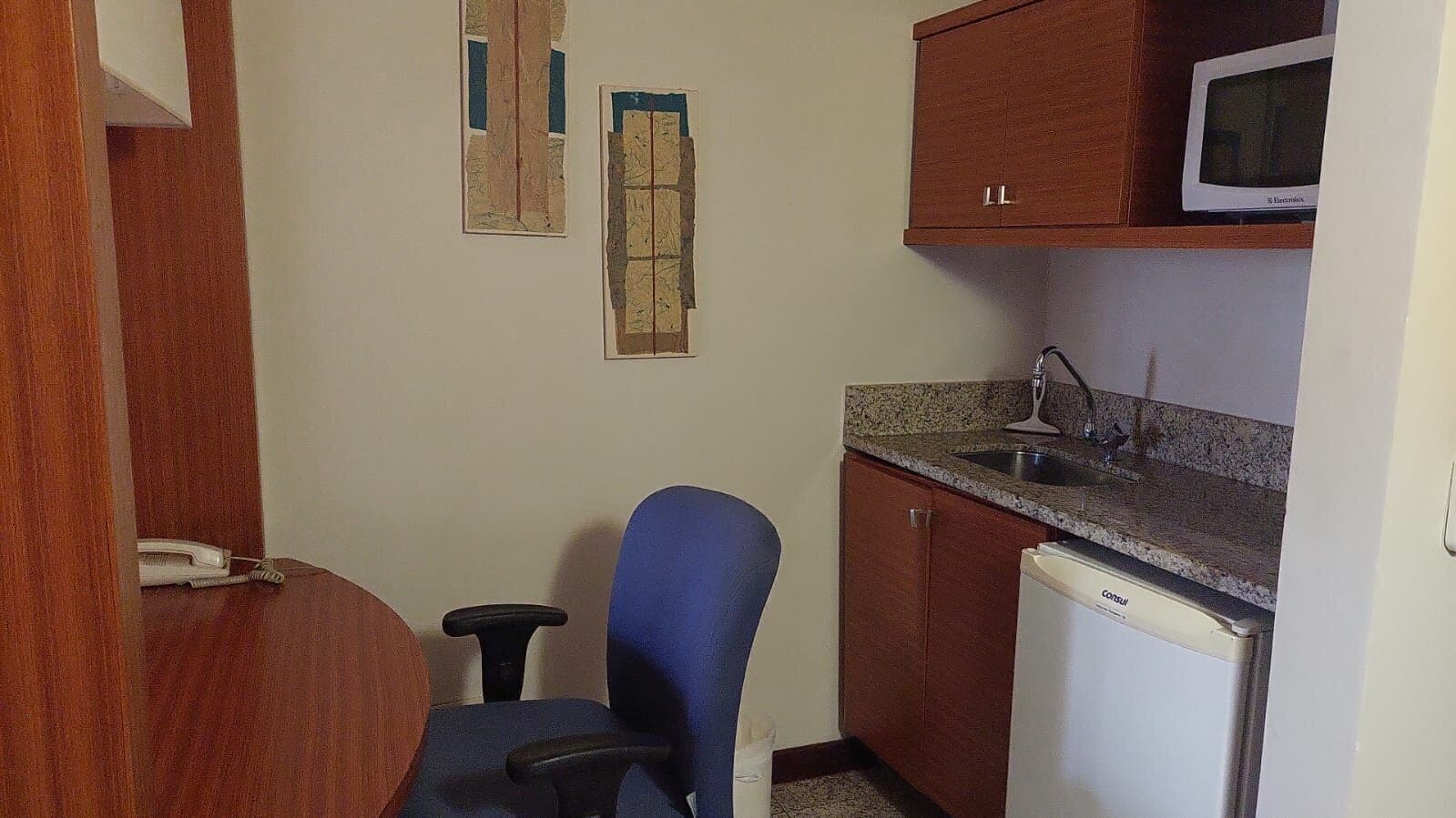 Une cuisine compacte équipée d’un four à micro-ondes, d’un évier et d’un réfrigérateur. On y trouve une chaise bleue et une petite table avec un téléphone.