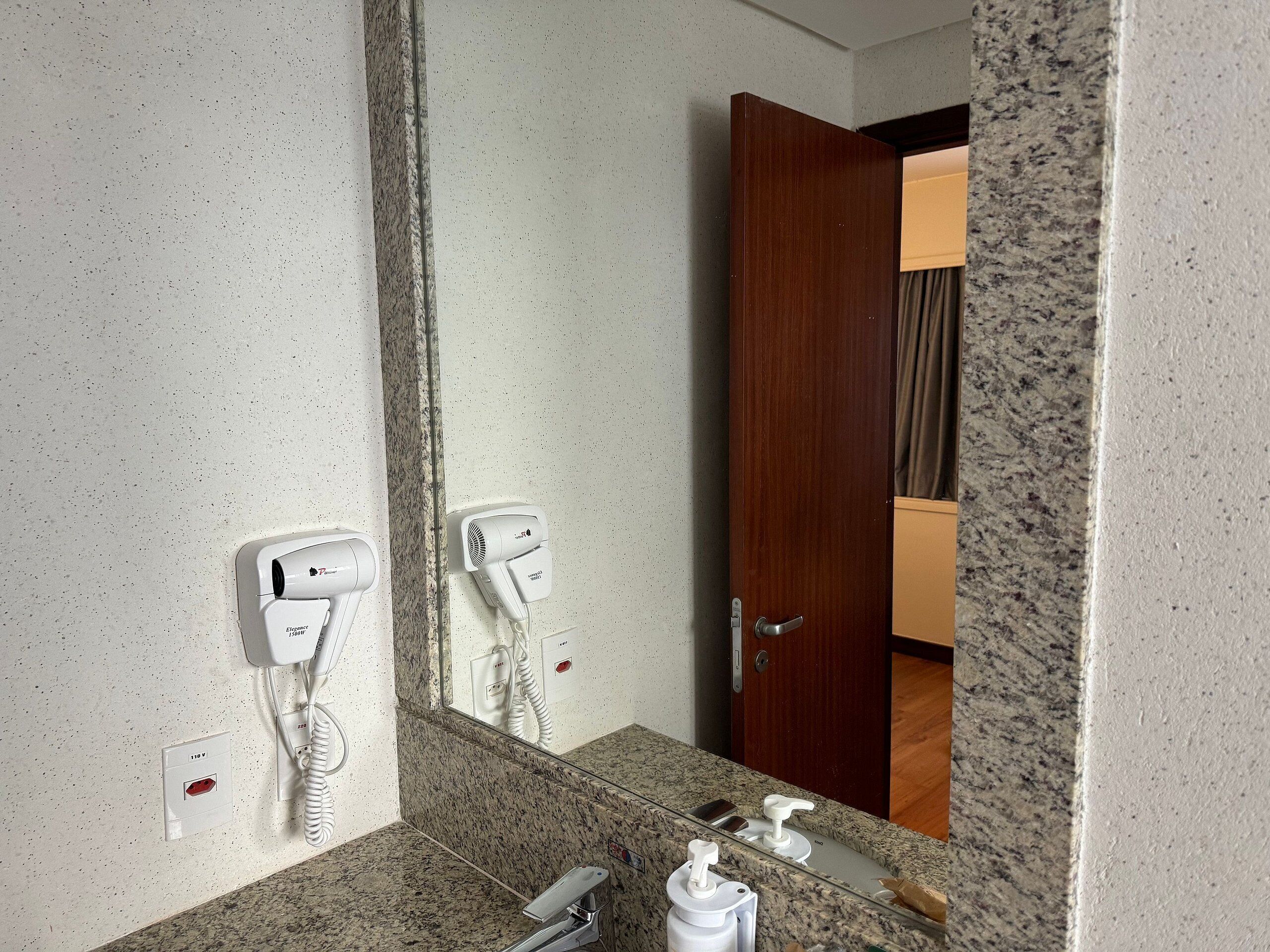 A bathroom with a granite countertop, a mirror, a hairdryer, and a towel dispenser.