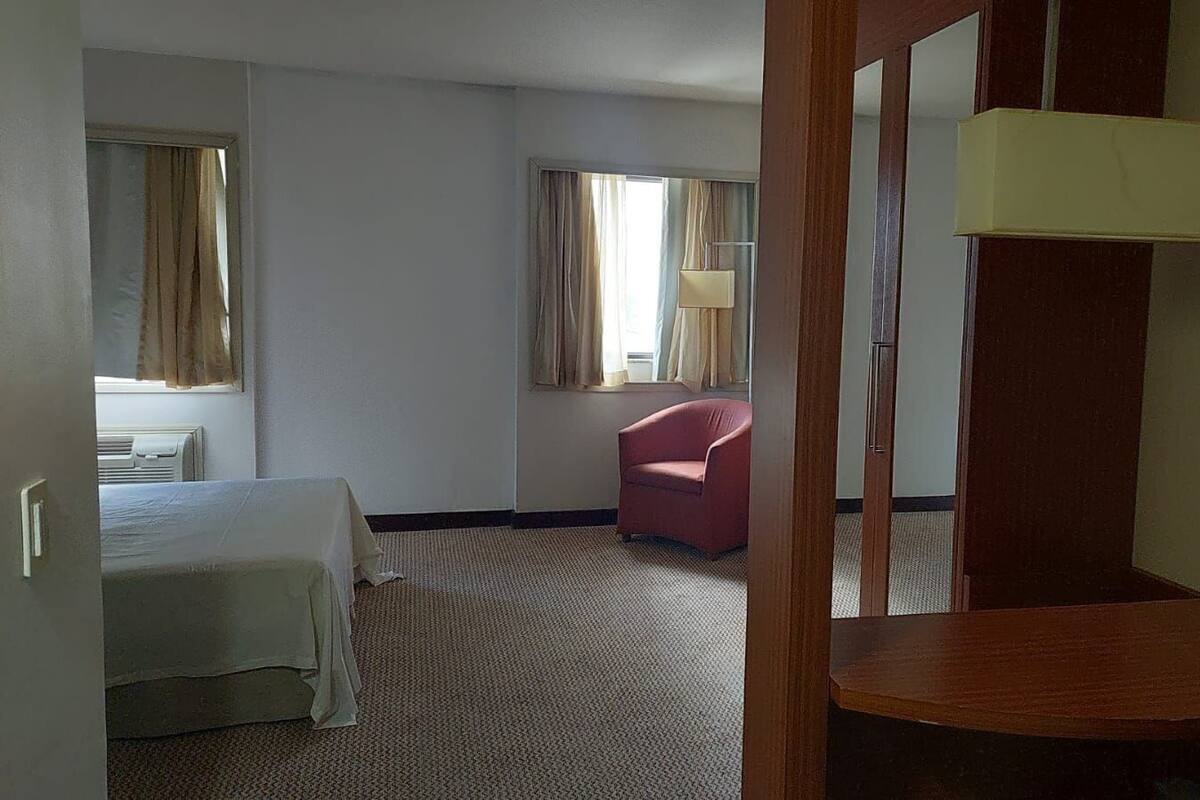 A hotel room with a bed, a red chair, a wooden desk, a telephone, and a window with curtains.