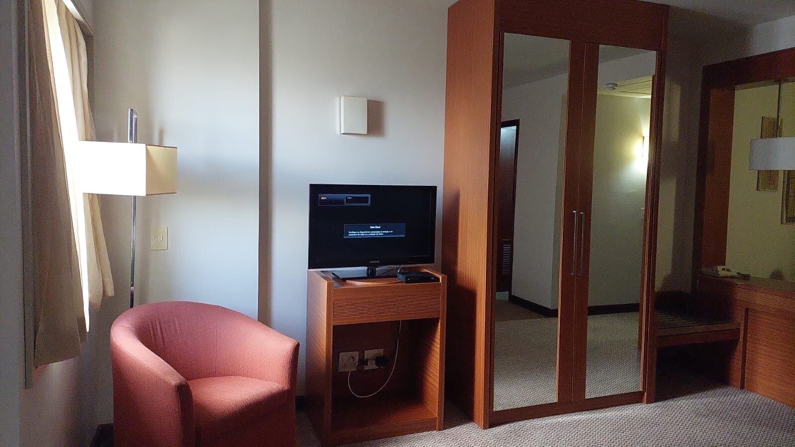 A hotel room with a television on a wooden stand, a red armchair, a wardrobe, and a mirror.