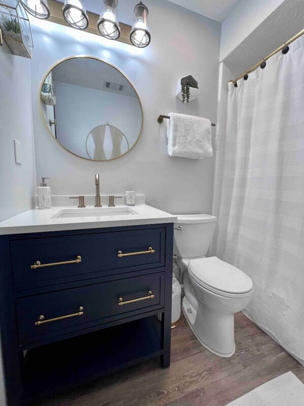 Un baño con tocador azul oscuro, un lavabo blanco, un espejo redondo, un inodoro y una cortina de ducha.