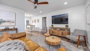 Una sala de estar moderna con un sofá de cuero, una mesa de centro de madera y un sillón gris. Se observa un aparador de madera con plantas y un televisor de pantalla plana montado en la pared.