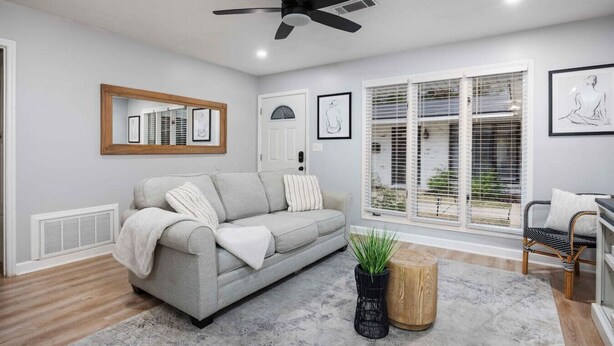 A modern living room with a grey sofa, a ceiling fan, a large window with blinds, and a framed artwork on the wall.