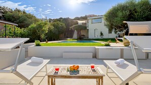 Zona de piscina con sillas de descanso, una mesa con frutas y una bebida, y vistas a una casa moderna y un jardín.