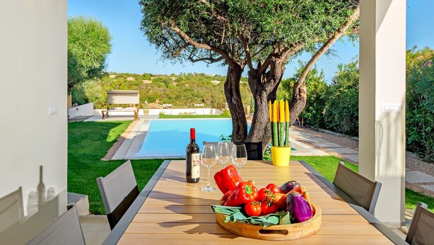 Una mesa junto a la piscina con una botella de vino, copas y un bol de verduras.