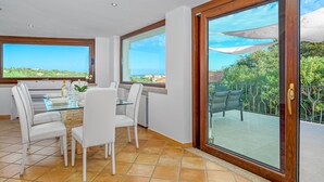 Un área de comedor con una mesa de vidrio y sillas blancas, ventanas amplias con vistas a zonas verdes y edificios, y un gran ventanal corredizo que da a un balcón exterior.