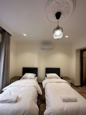 A hotel room with two beds, a ceiling light, an air conditioning unit, and a window with curtains. - 3 Antica Jabalal-lwebdeh (Amman)