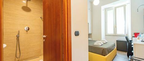 A bathroom with a shower, a toilet, and a sink. There is a bed with a yellow mattress and a desk with a chair.