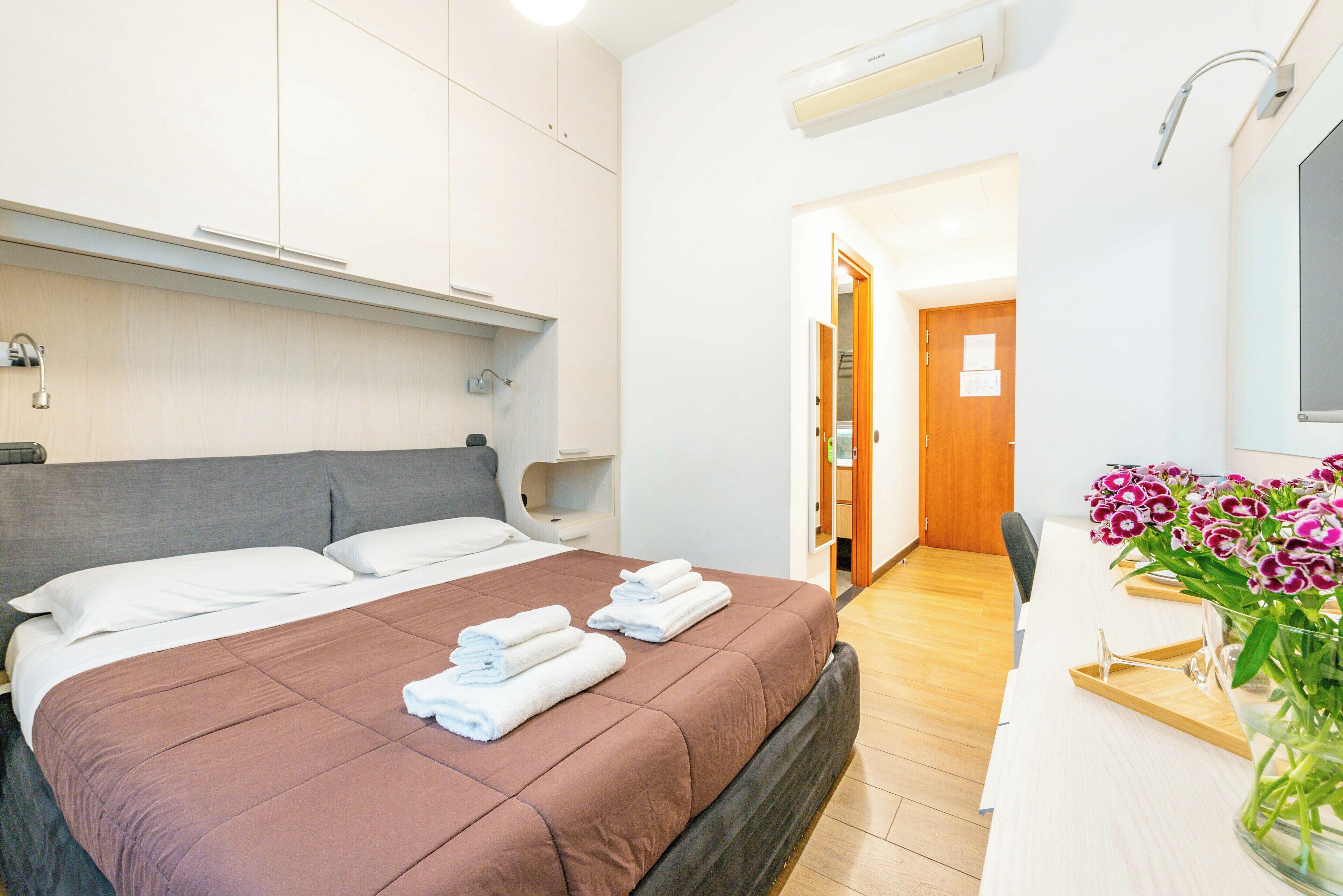 Ein Hotelzimmer mit Bett, Nachttischen, einem Schreibtisch mit Stuhl und einer Blumenvase.