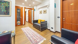 A hotel lobby with a reception desk, a sofa with yellow cushions, a patterned rug, and a wooden door with a number plaque. - Andenis Tiber House  (Rome)