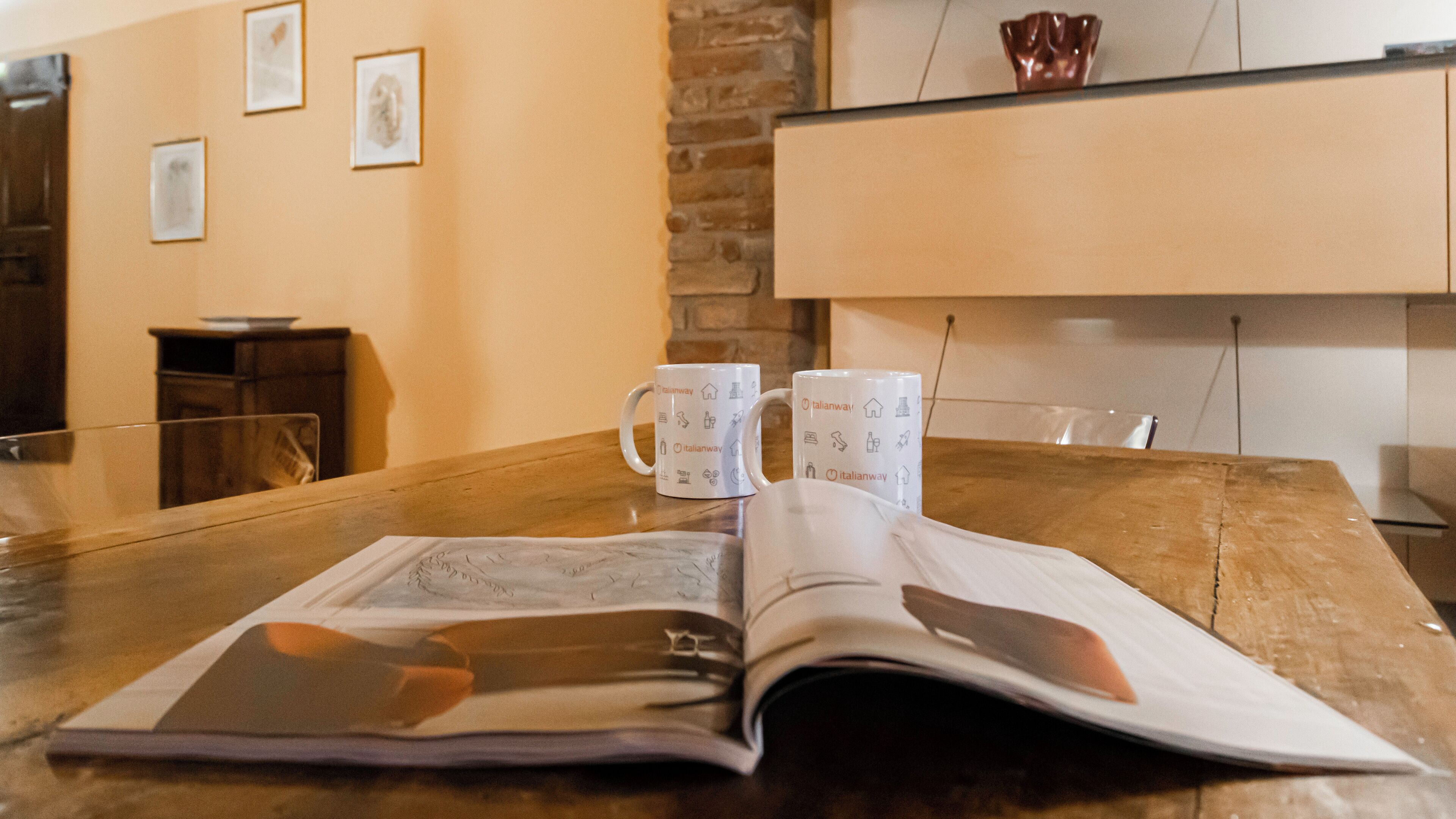 An open magazine on a wooden table with two white mugs featuring Italianway logos.