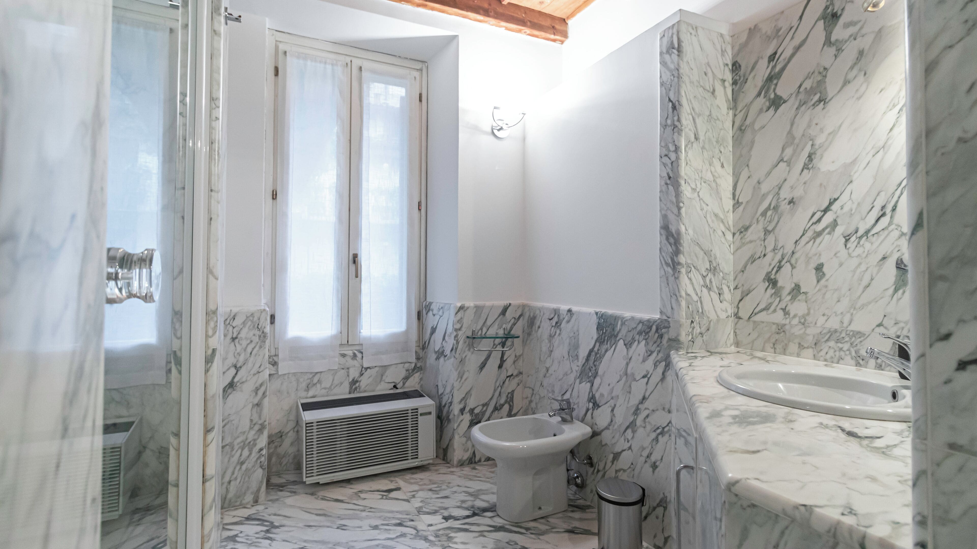 A bathroom with a marble floor, a bidet, a sink, and a window with curtains.