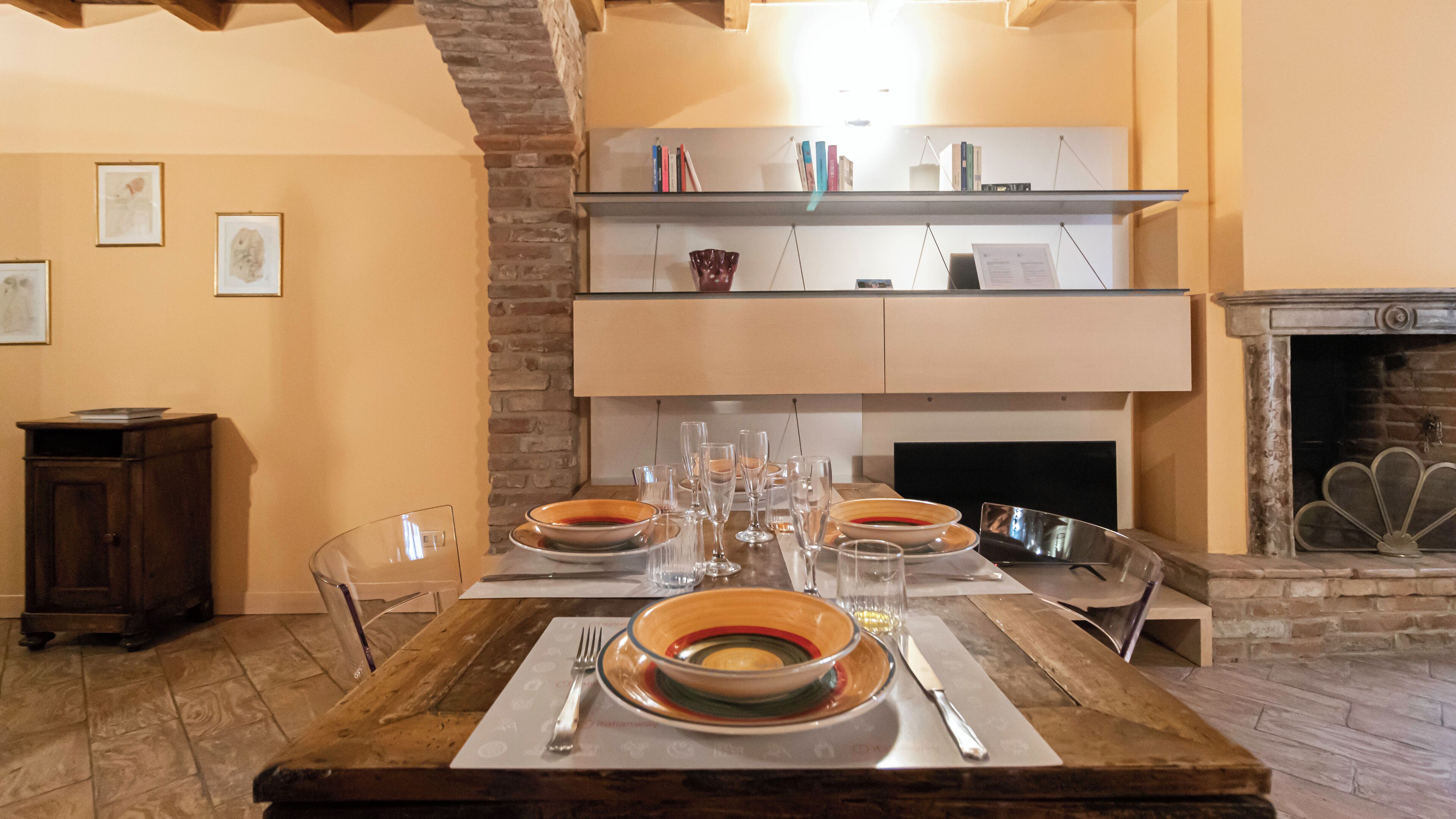 A dining area with a long table set for four, surrounded by chairs. There is a fireplace with a stone mantle and a bookshelf above it.
