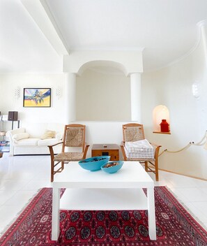 A modern living room with a white sofa, two wooden chairs, a white coffee table with blue bowls, a red patterned rug, and a painting on the wall. - 54 Golfmar Carvoeiro apartment with fabulous pool (Carvoeiro)