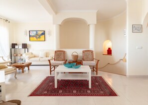 Een ruime woonkamer met een witte bank, twee fauteuils, een salontafel en een rood tapijt met een patroon. Je ziet een boekenkast met boeken en speakers, een schilderij aan de muur en een kleine tafel met een lamp.