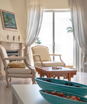 Sala de estar com duas poltronas, uma mesa de centro, lareira e vista para palmeiras através de uma porta de vidro.