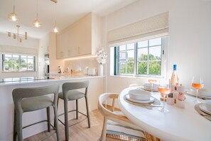 A modern kitchen with a dining area, featuring a central island, bar stools, and a view through large windows.
