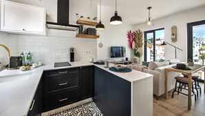 Een moderne keuken met witte en zwarte kastjes, een inbouwoven en een groot eiland. Er is een eethoek met een houten tafel en stoelen, en een groot raam met uitzicht op de stad.