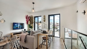 Een moderne woonkamer met een bank, een eettafel en een televisie. Er zijn grote ramen en een balkon.