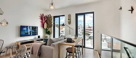 A modern living room with a sofa, a dining table, and a TV. There are large windows and a balcony.
