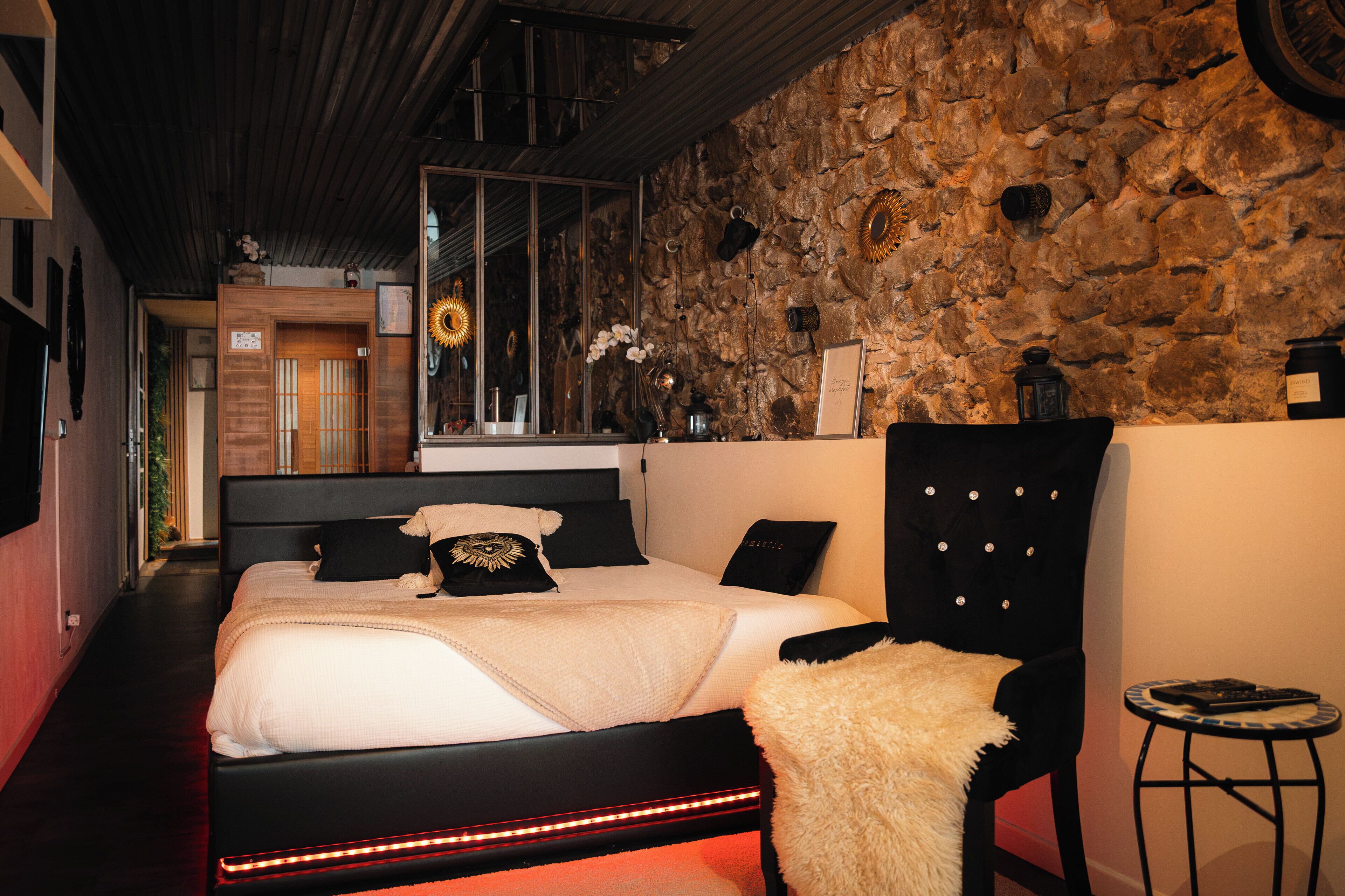 Un dormitorio con pared de piedra, una cama con ropa de cama blanco y negro, una silla negra y una mesita de noche con lámpara.
