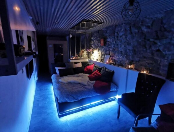 Une chambre moderne avec un mur en pierre, un lit doté d’un éclairage bleu sous le lit et une table de chevet avec des bougies.