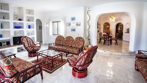 A spacious living room with a sofa set, armchairs, and a coffee table. There is a television on a stand and a bookshelf with various decorative items.