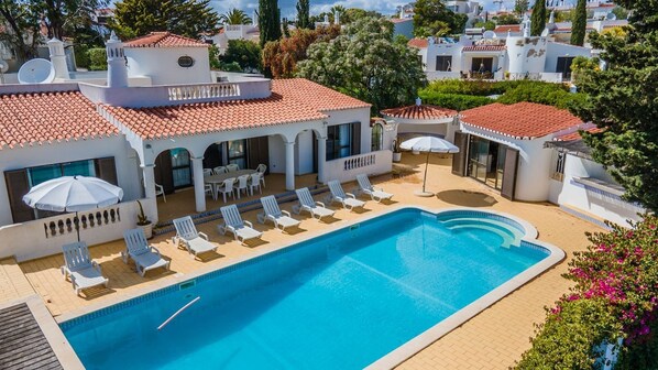 Relax by the clear blue pool surrounded by lounge chairs and umbrellas, adjacent to a Mediterranean-style building with a covered patio.
