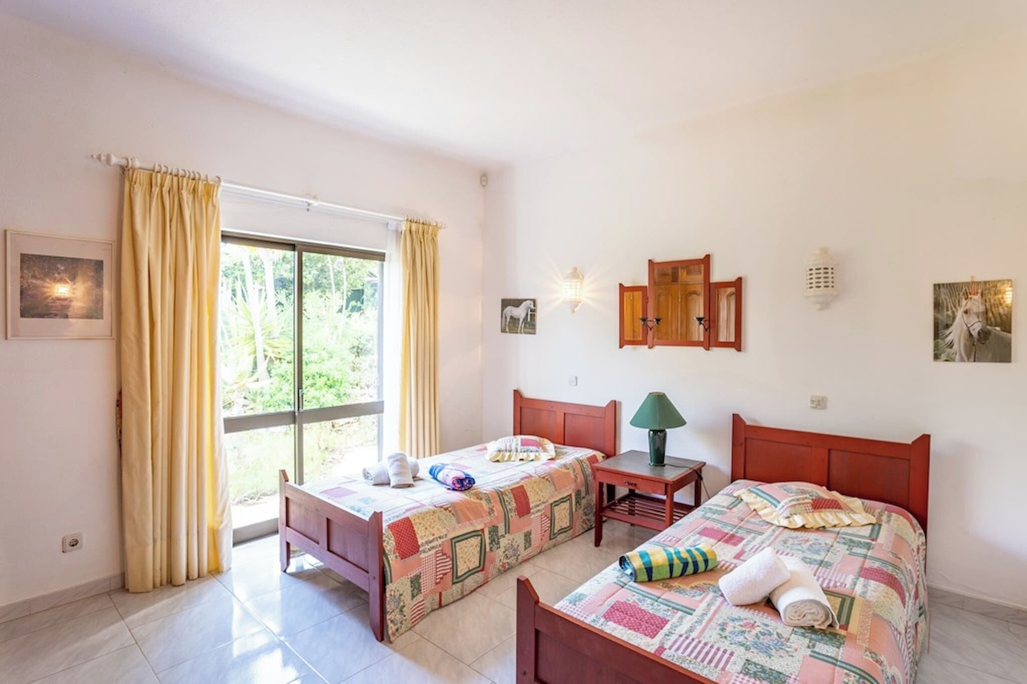 A room with two beds, a window with curtains, a small table with a lamp, and wall-mounted lights.