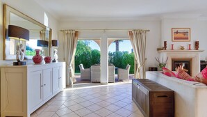 A spacious living room with a white sofa, a coffee table, and a fireplace. There is a dining area with wicker furniture and a view of greenery outside.