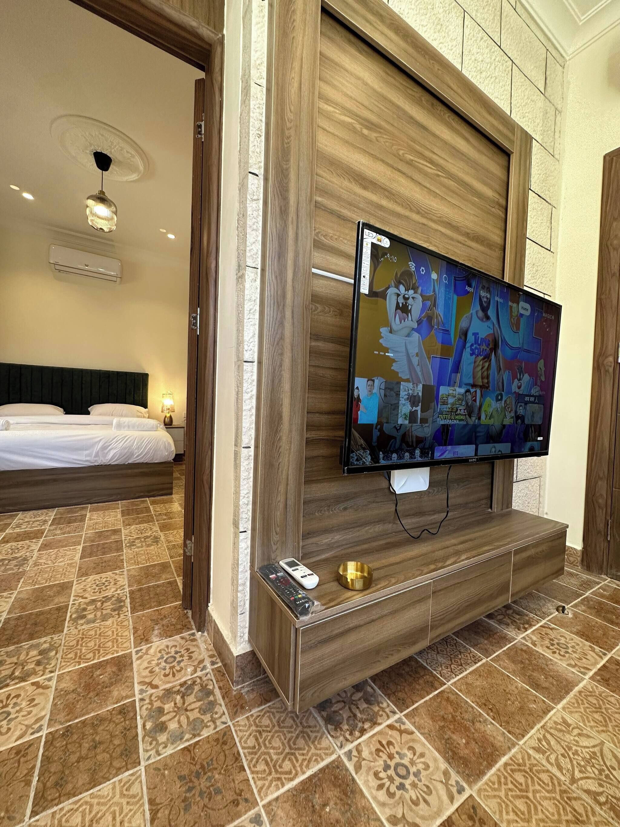 A hotel room with a large flat-screen TV mounted on a wooden wall, a bed with white linens, patterned floor tiles, and a small table with a remote control and a decorative bowl.