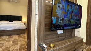 A hotel room with a large flat-screen TV mounted on a wooden wall, a bed with white linens, patterned floor tiles, and a small table with a remote control and a decorative bowl. - Antica Jabalal-lwebdeh (Amman)