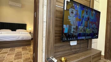 A hotel room with a large flat-screen TV mounted on a wooden wall, a bed with white linens, patterned floor tiles, and a small table with a remote control and a decorative bowl.