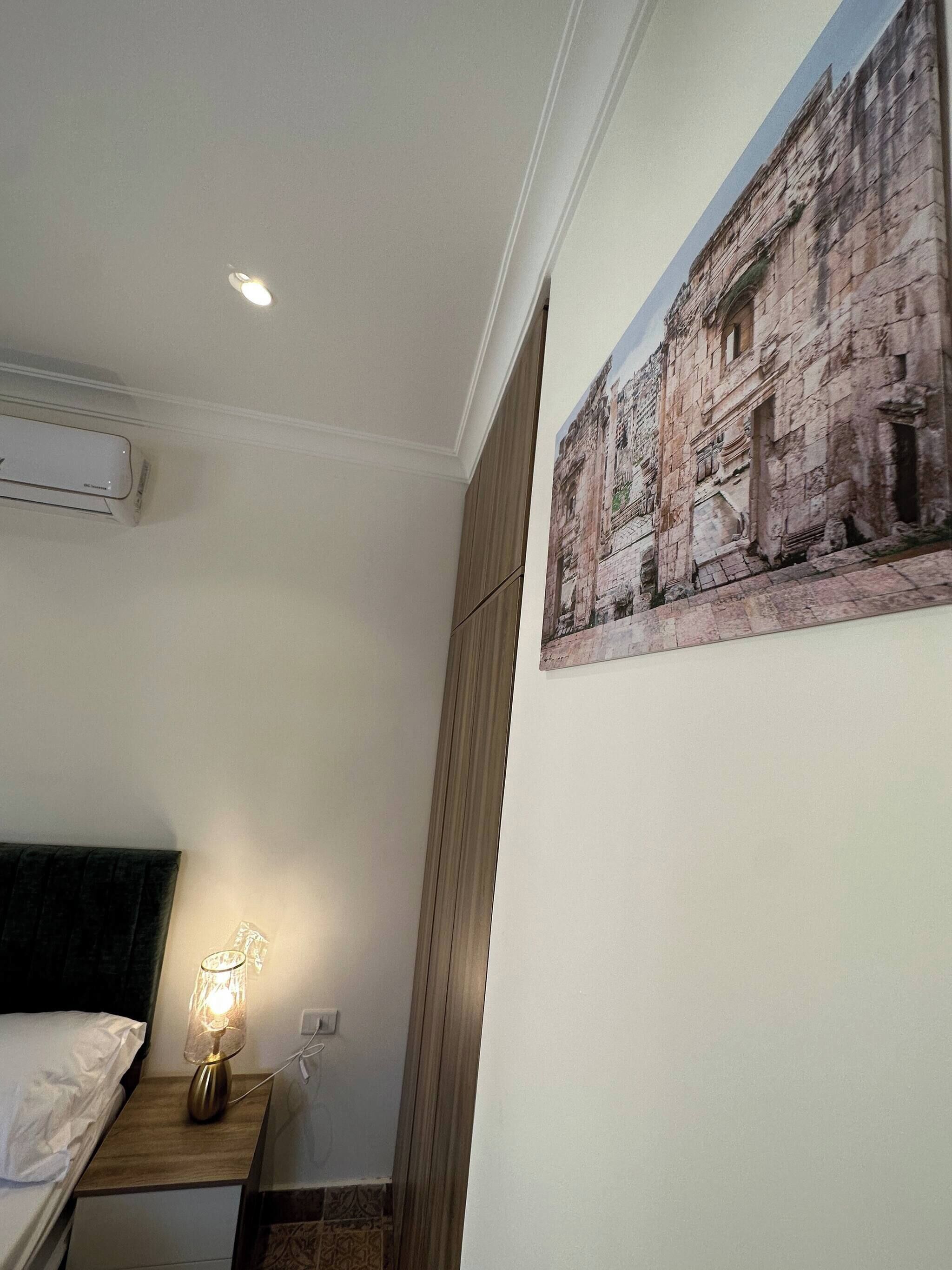 A bedroom with a bed, bedside table with a lamp, an air conditioning unit, a painting on the wall, and a wooden wardrobe.