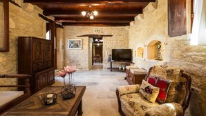 A stone-walled room with a wooden ceiling, a television, a sofa, and a dining area in the background. - Ani Villa, authentic Cretan lifestyle (Rethymno)