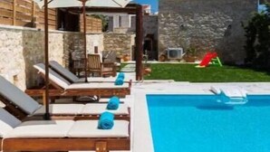 A pool area with lounge chairs and umbrellas, a stone building in the background, and a clear blue sky. - Ani Villa, authentic Cretan lifestyle (Rethymno)