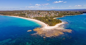 Vue aérienne d’une ville côtière avec une plage, des eaux d’un bleu limpide et une zone boisée en arrière-plan.