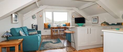 A cozy attic room with a teal sofa, wooden flooring, a dining area with a table and chairs, and a kitchen with white cabinets and a TV.