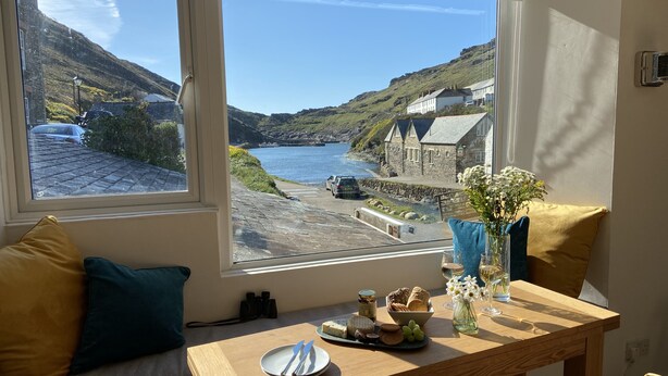 Quarto com uma grande janela com vista para a área costeira, uma mesa de madeira com um prato, talheres e um vaso com flores.