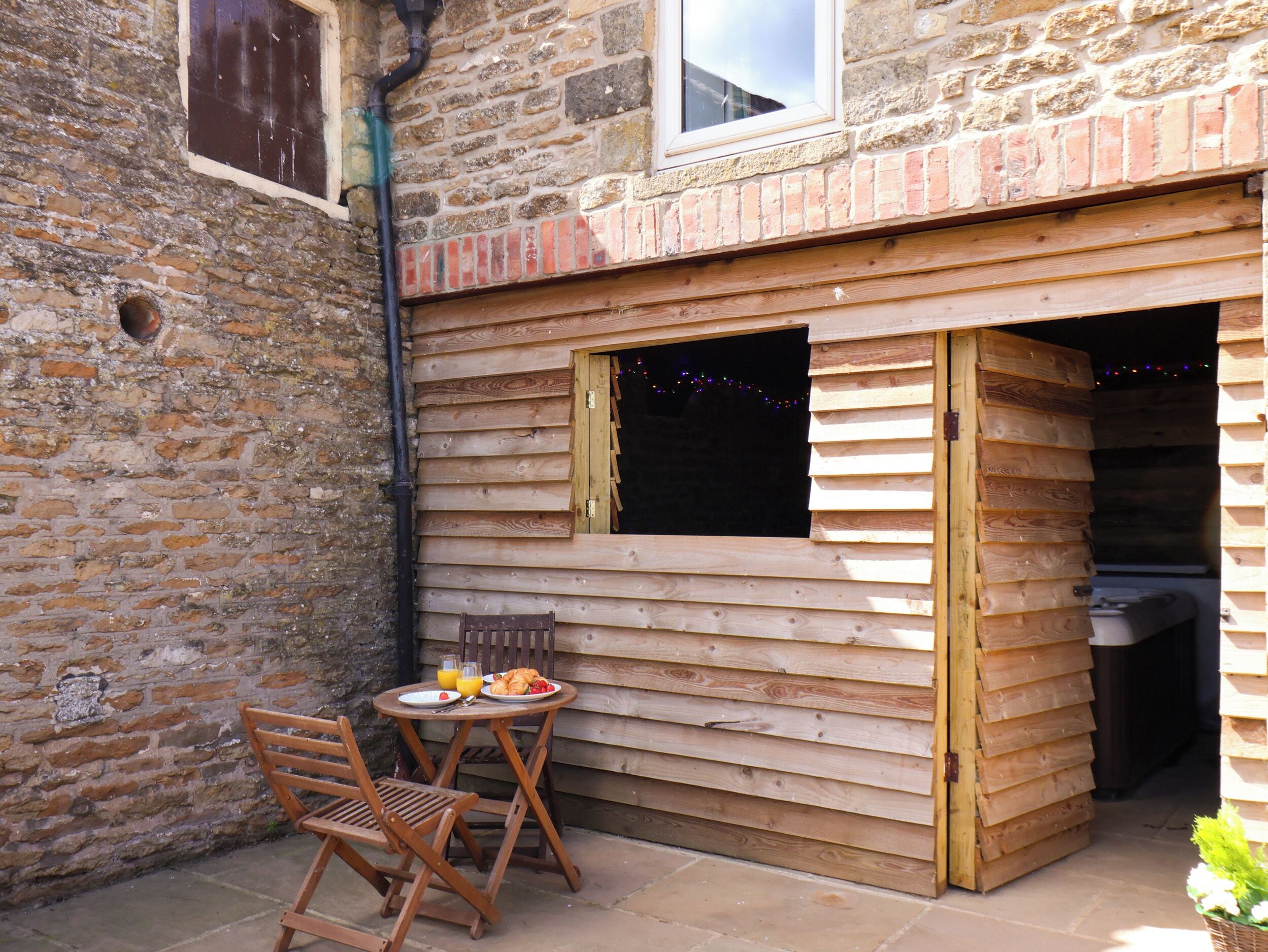 A wooden outdoor seating area with a table and chairs, a brick wall, and a window.