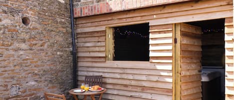 A wooden outdoor seating area with a table and chairs, a brick wall, and a window.