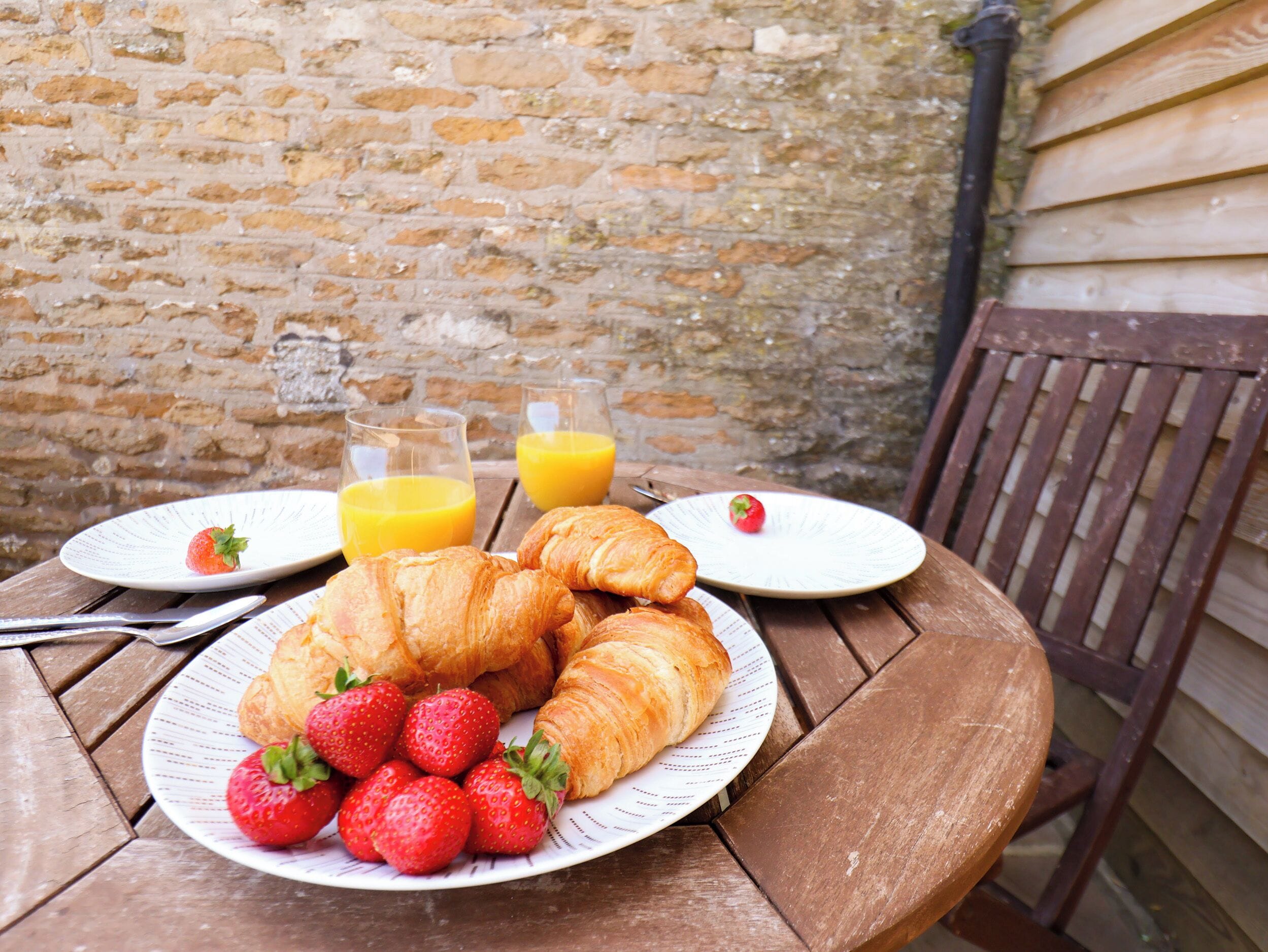 Un tavolo di legno con piatti di croissant e fragole, due bicchieri di succo d'arancia e un muro di mattoni sullo sfondo.