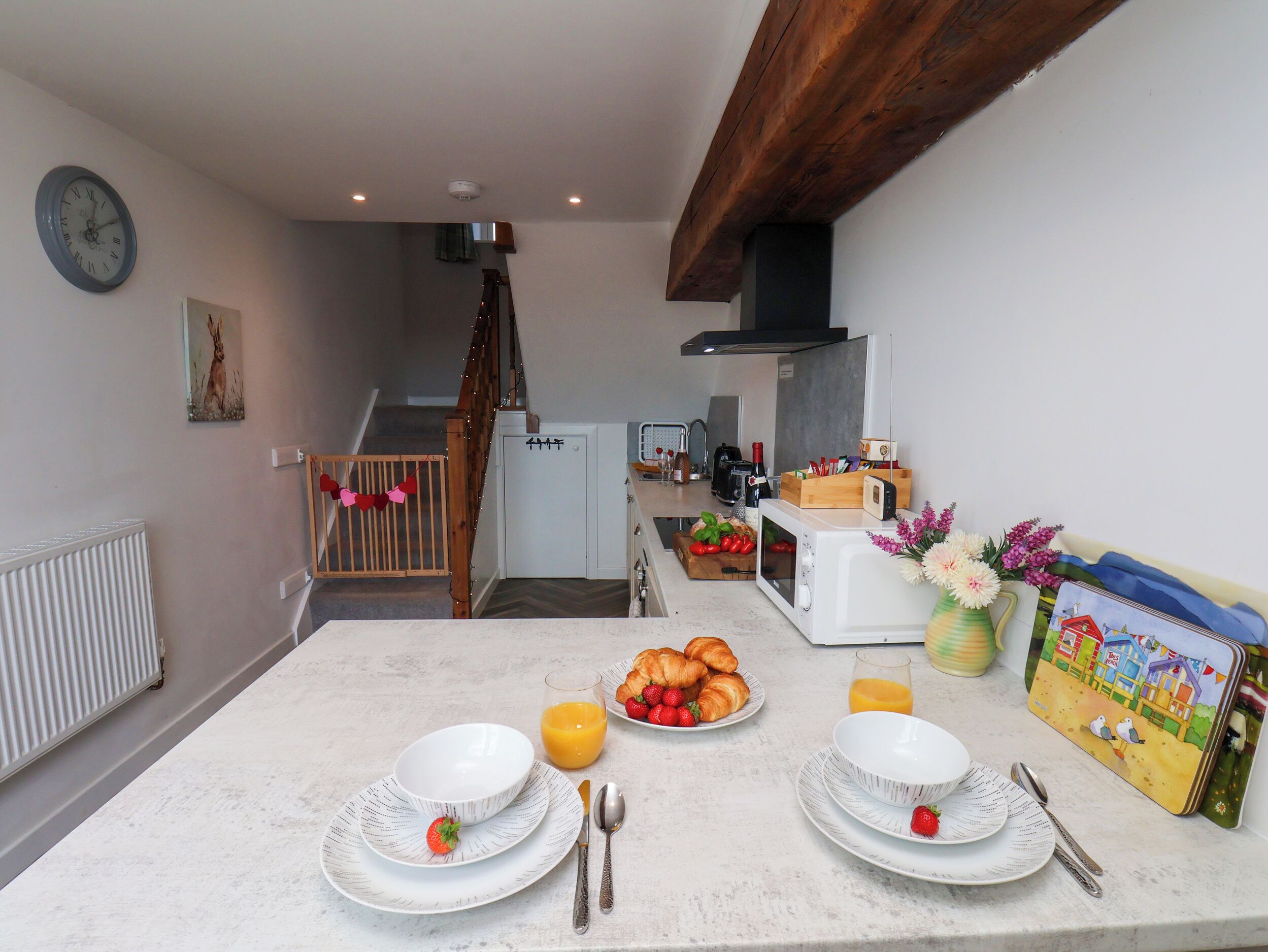Una cocina con una mesa de comedor preparada para el desayuno, un reloj en la pared y una escalera que lleva a otra habitación.