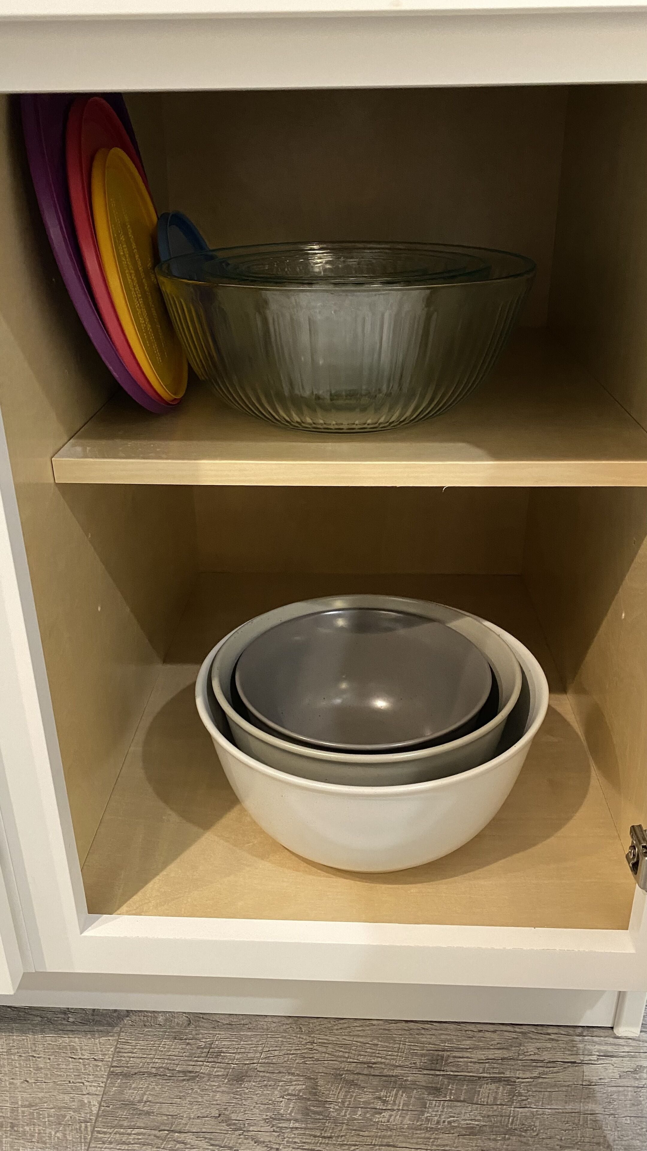 Un mueble de cocina con dos estantes. El estante superior sostiene un cuenco de vidrio transparente y tapas de plástico de colores. El estante inferior contiene un cuenco blanco con tapa.