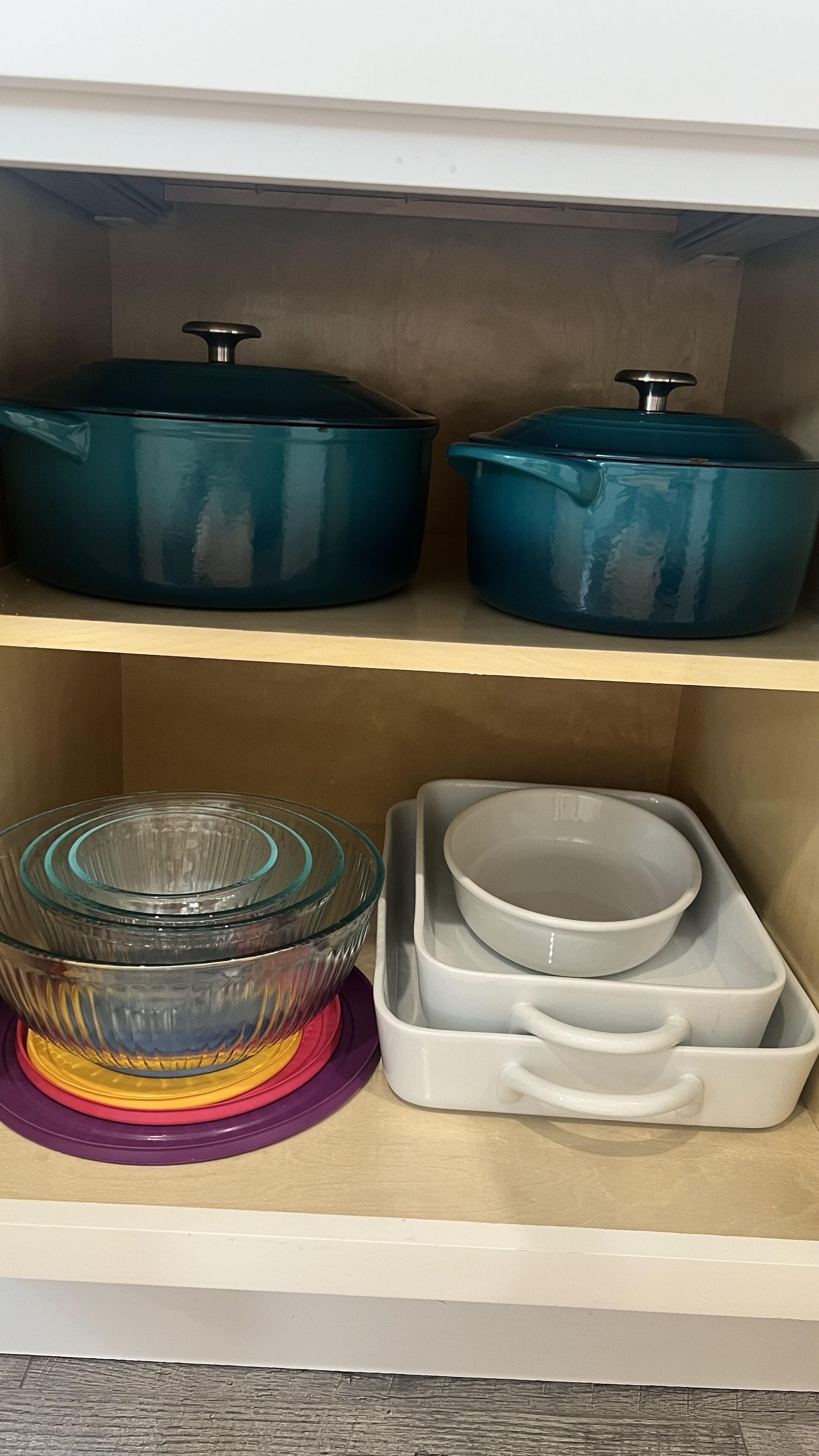 A kitchen shelf with two teal-colored pots, a glass bowl on a purple base, and a white dish rack with bowls.