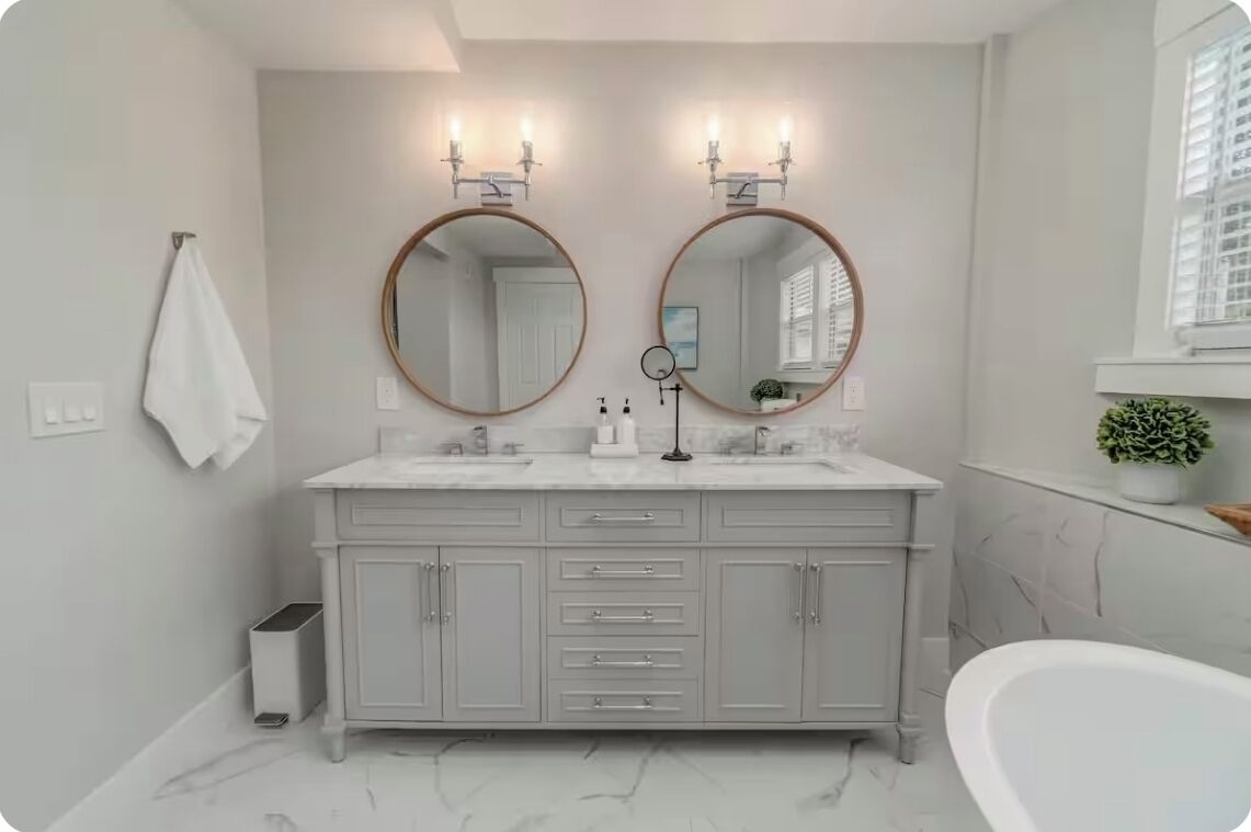 Une salle de bain avec un meuble-lavabo blanc, deux miroirs ronds, une baignoire blanche et une petite plante en pot posée sur une étagère.