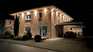 A modern two-story house with a garage, illuminated exterior lights, and a well-maintained lawn. - Boardinghouse - Apartment (Lippstadt)