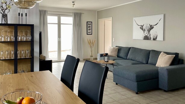 A modern living room with a grey sectional sofa, a dining table with chairs, and a glass cabinet with shelves. - Boardinghouse - Apartment (Lippstadt)