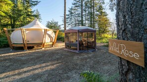 Een kampeerterrein met tenten, een tafel en stoelen, een bordje met de tekst 'Wild Rose' en een bebost landschap op de achtergrond.